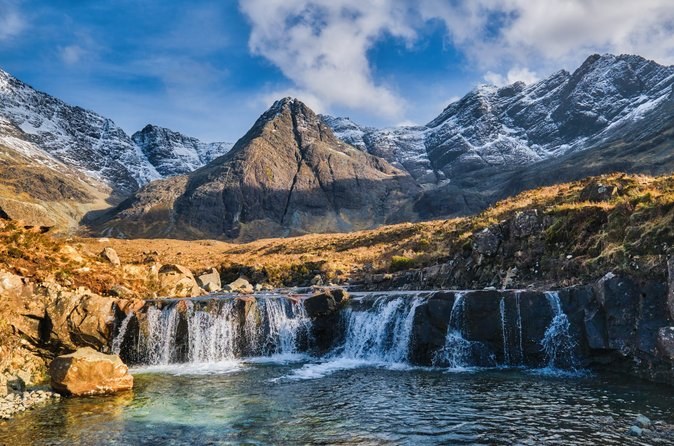 The Fairy Pools falls in autumn, rust-coloured grass on the banks and snow on the Cuillin ridge above
