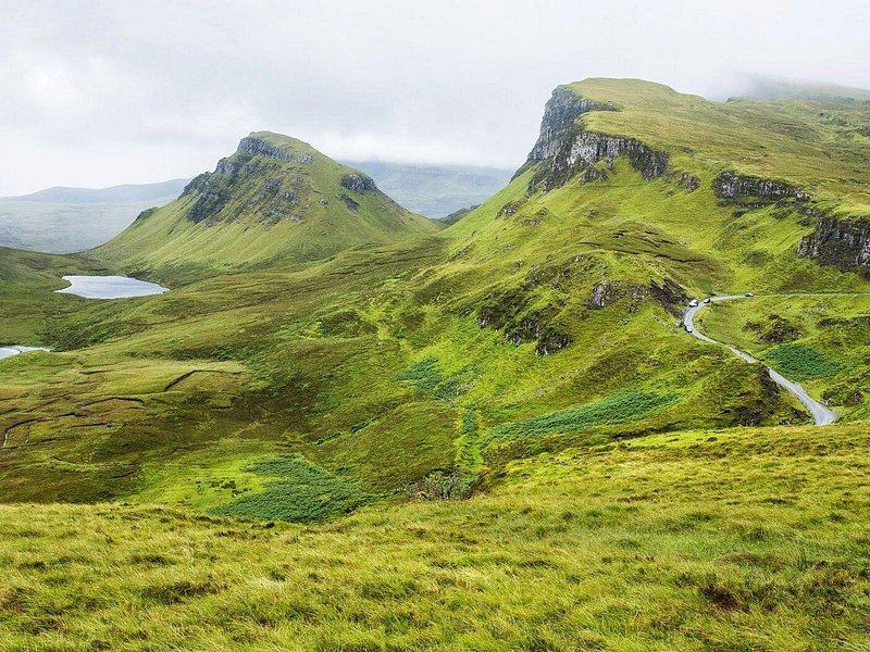 The Quiraing in summer, steep green slopes and a flat-topped ridge above a lochan, the road running along the hillside