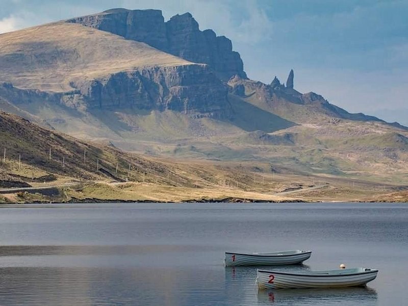 The Storr cliffs and the Old Man standing clear of them, seen across a still loch with two numbered rowing boats moored in the foreground